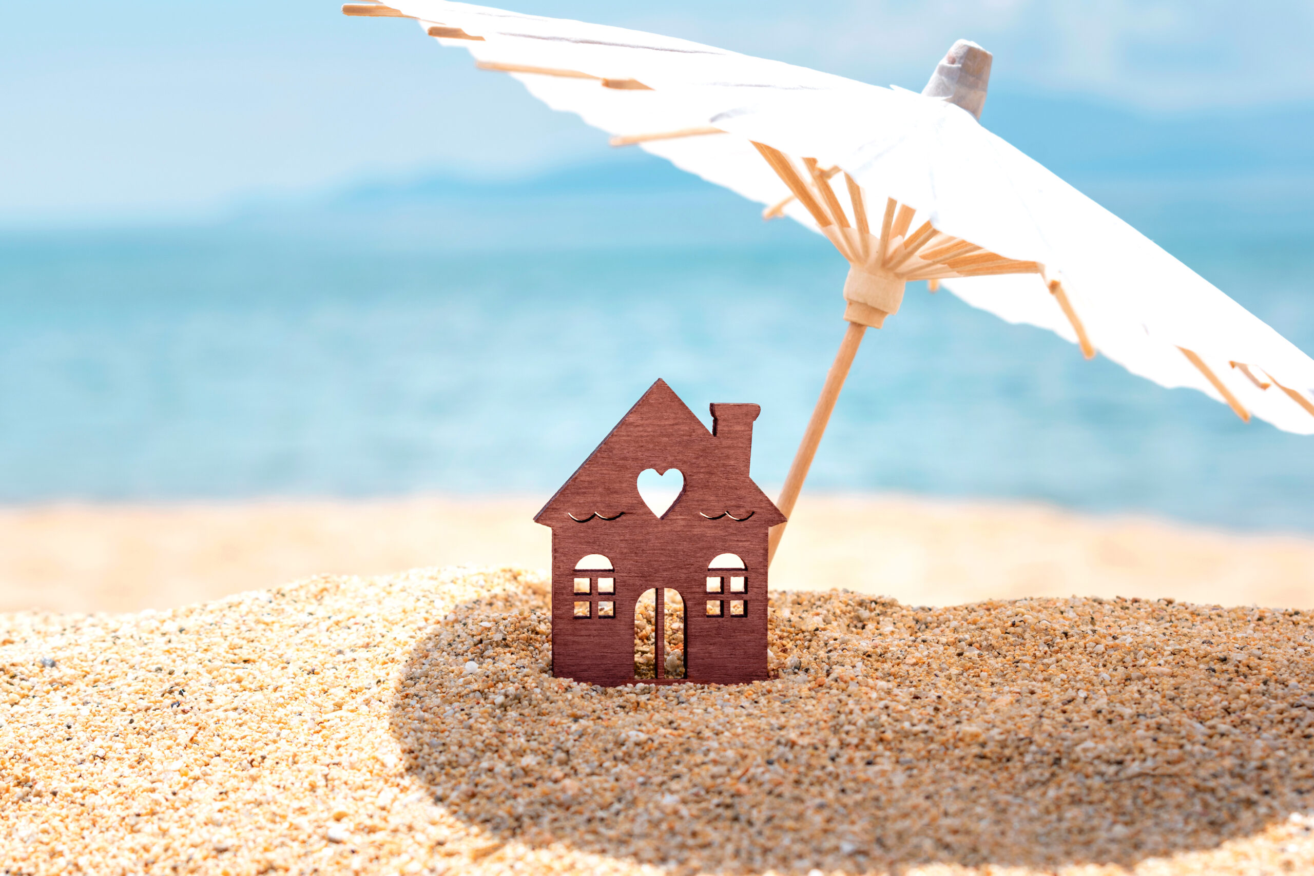 miniature house and umbrella on beach, blue sea and sky on blurred background. vacation home for happy holiday for family. real estate, sale or property investment concept. closeup. copy space